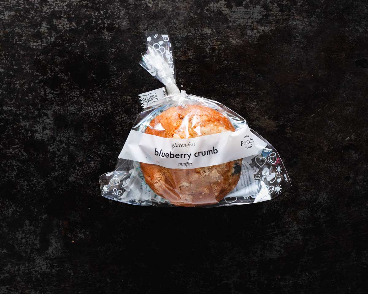 Packaged blueberry crumb muffin in clear packaging against a dark textured background.