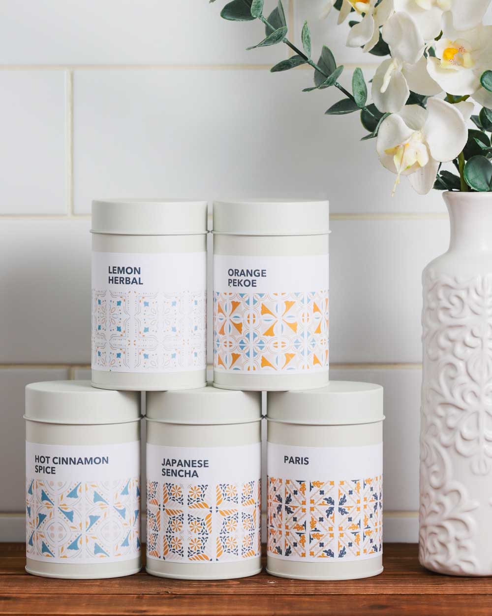 Four cylindrical tea tins with geometric patterns in white, orange,and bluecolors, labeled with different flavors such as Lemon Herbal, Orange Pekoe, Hot Cinnamon Spice, Japanese Sencha, and Paris, displayed on a kitchen counter.