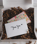 Valentine's gift box with a floral card and an envelope labeled 'For You' on top of brown shredded paper.