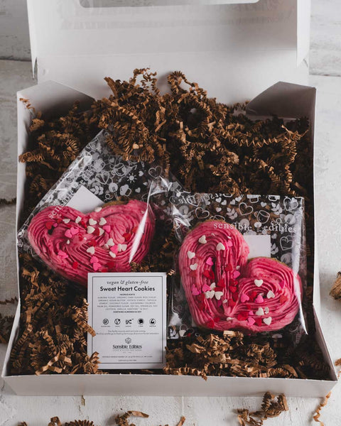 Valentine's gift box containing two heart-shaped cookies with pink frosting, with a label for 'Sensible Edibles Sweet Heart Cookies' on top of brown shredded paper.