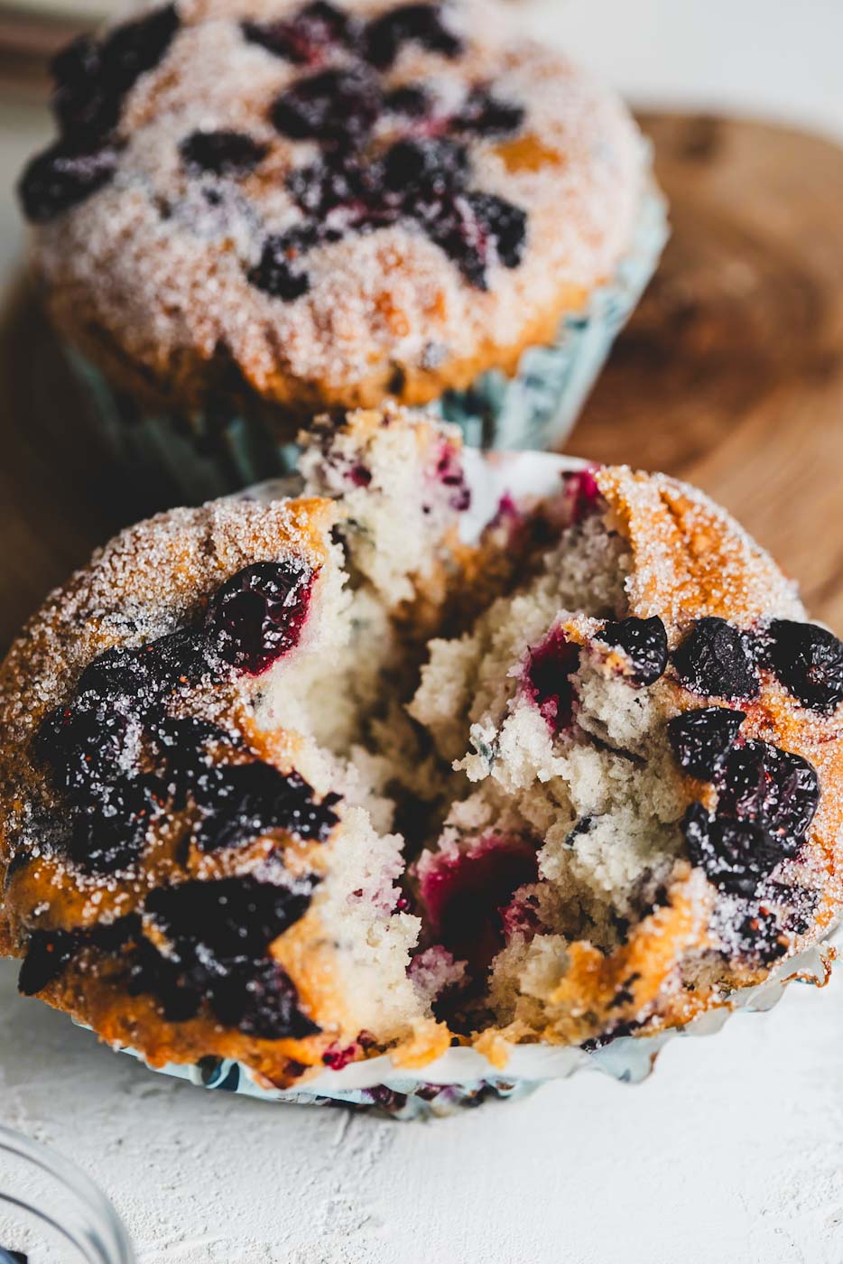Vanilla berry muffin broken open to reveal a moist interior with visible berries, sprinkled with powdered sugar, on a wooden board.
