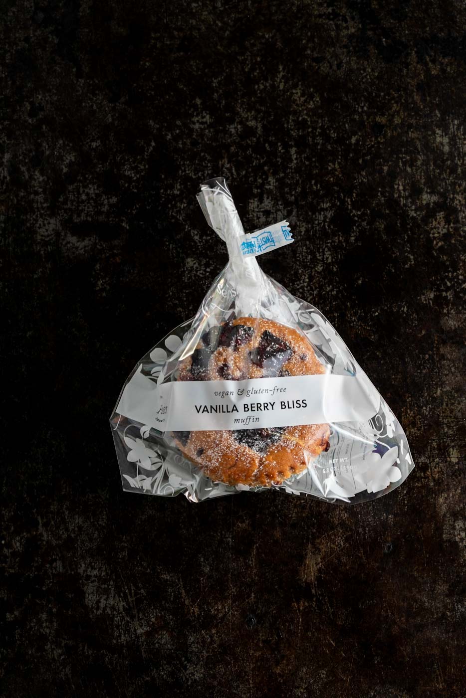 Packaged vanilla berry muffin labeled 'Vanilla Berry Bliss' in a clear bag with a floral design, against a dark background.