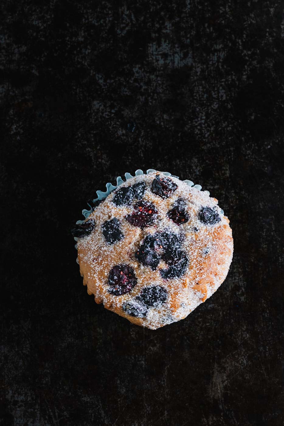 Close-up of a vanilla berry muffin topped with mixed berries and powdered sugar, against a dark background.