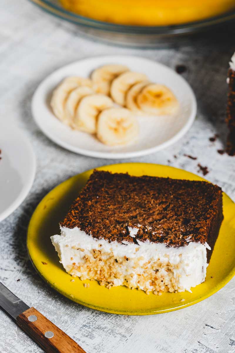 slice of banana walnut loaf with frosting and walnuts on top with sliced bananas in the background