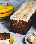 A banana walnut loaf cake with a slice cut out, alongside fresh bananas and walnuts on a grey background.