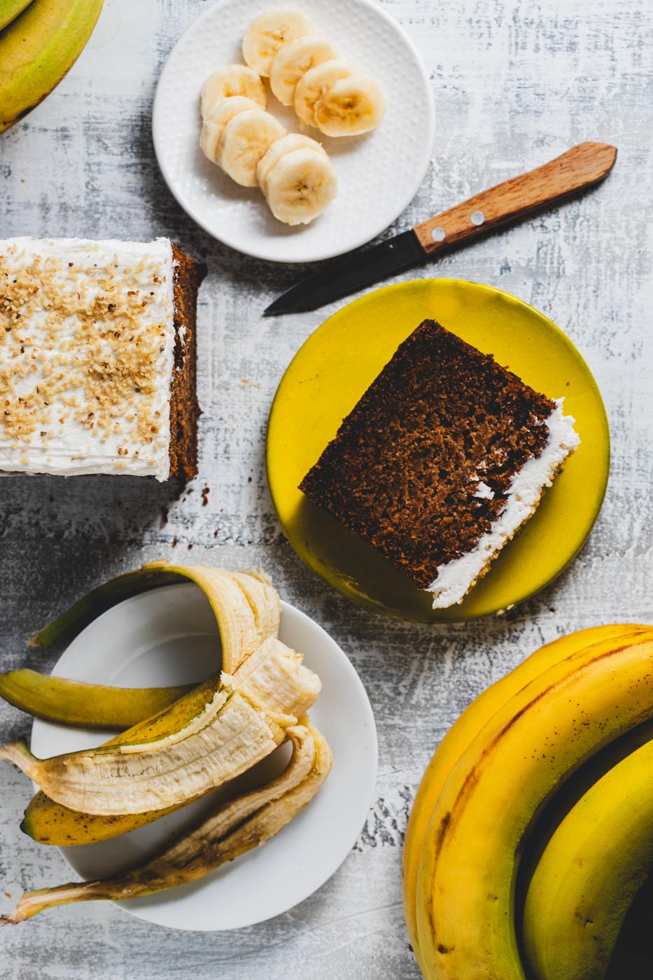 A banana walnut loaf cake with a slice cut out, alongside fresh bananas and walnuts on a grey background.
