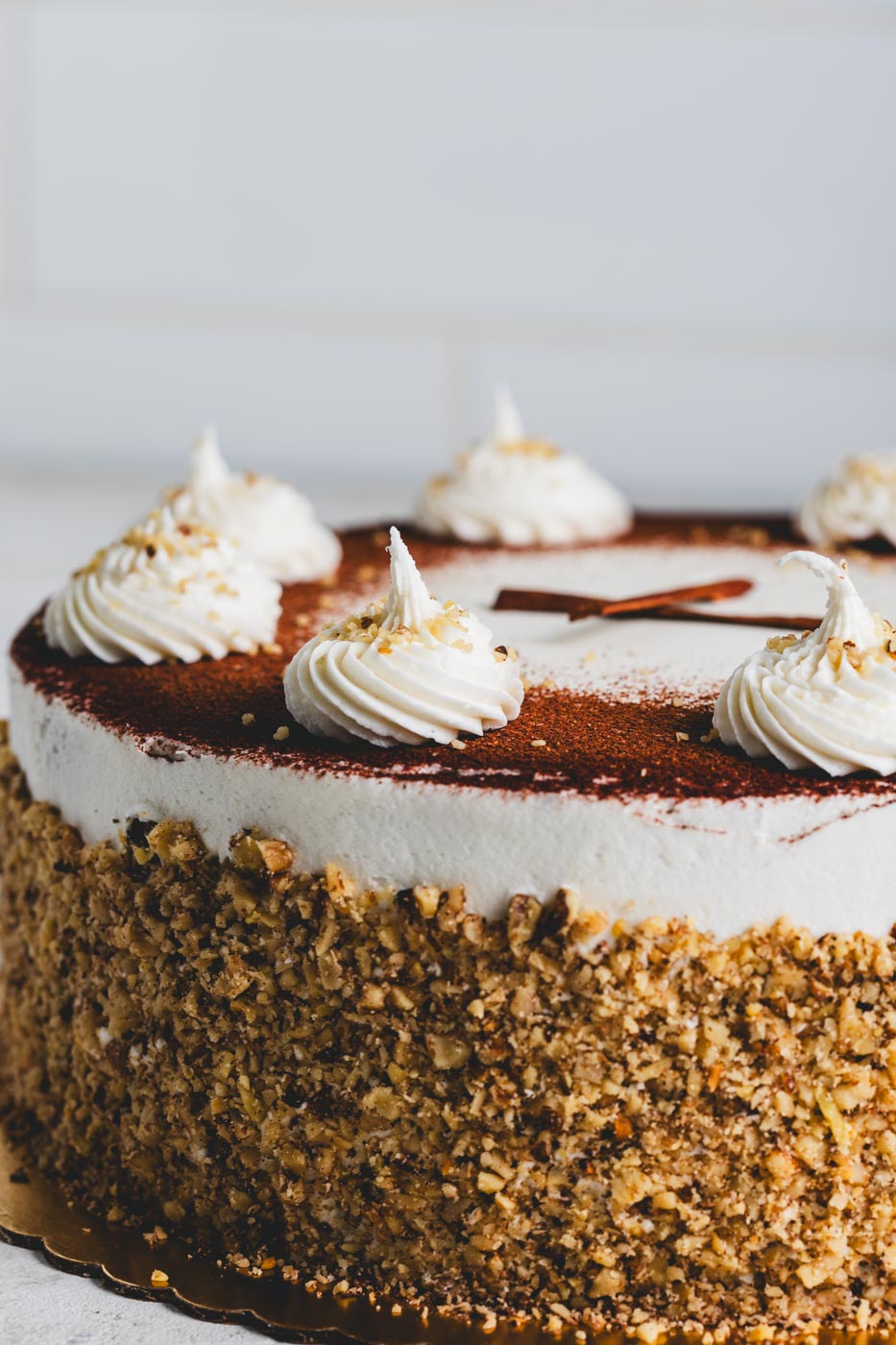 A close up of a cake with white frosting dusted with cooca powder and rolled in crushed nuts with white icing and cinnamon stick decoration