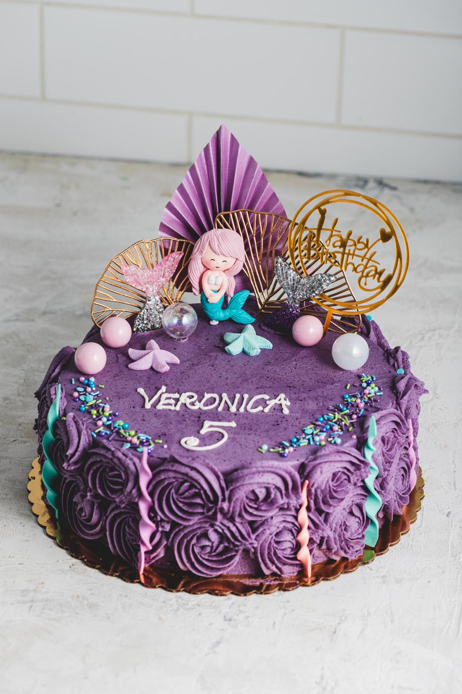 Cake with purple decorative icing and pink and blue mermaid themed decorations with 'veronica 5' written on top and a gold happy birthday decorative topper.
