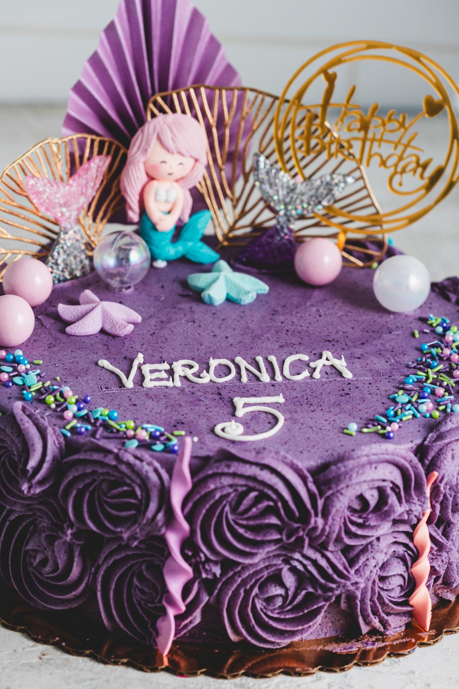Cake with purple decorative icing and mermaid themed decorations with 'veronica 5' written on top and a happy birthday decorative topper.