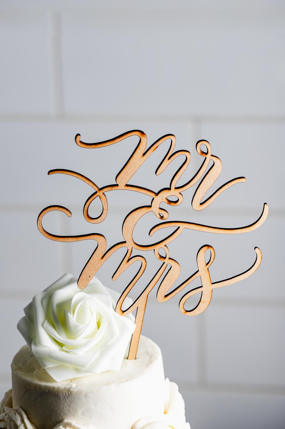 Close up of the top of a white tiered wedding cake with a white flower on top and a gold cake topper that says 