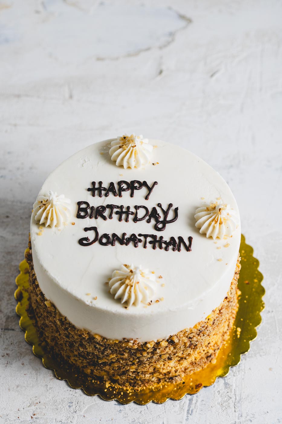 A vanilla frosted cake with "happy birthday jonathan" written in brown icing and decorative dollops of white icing topped with a crushed nut garnish. The side of the cake is rolled in crushed nuts.