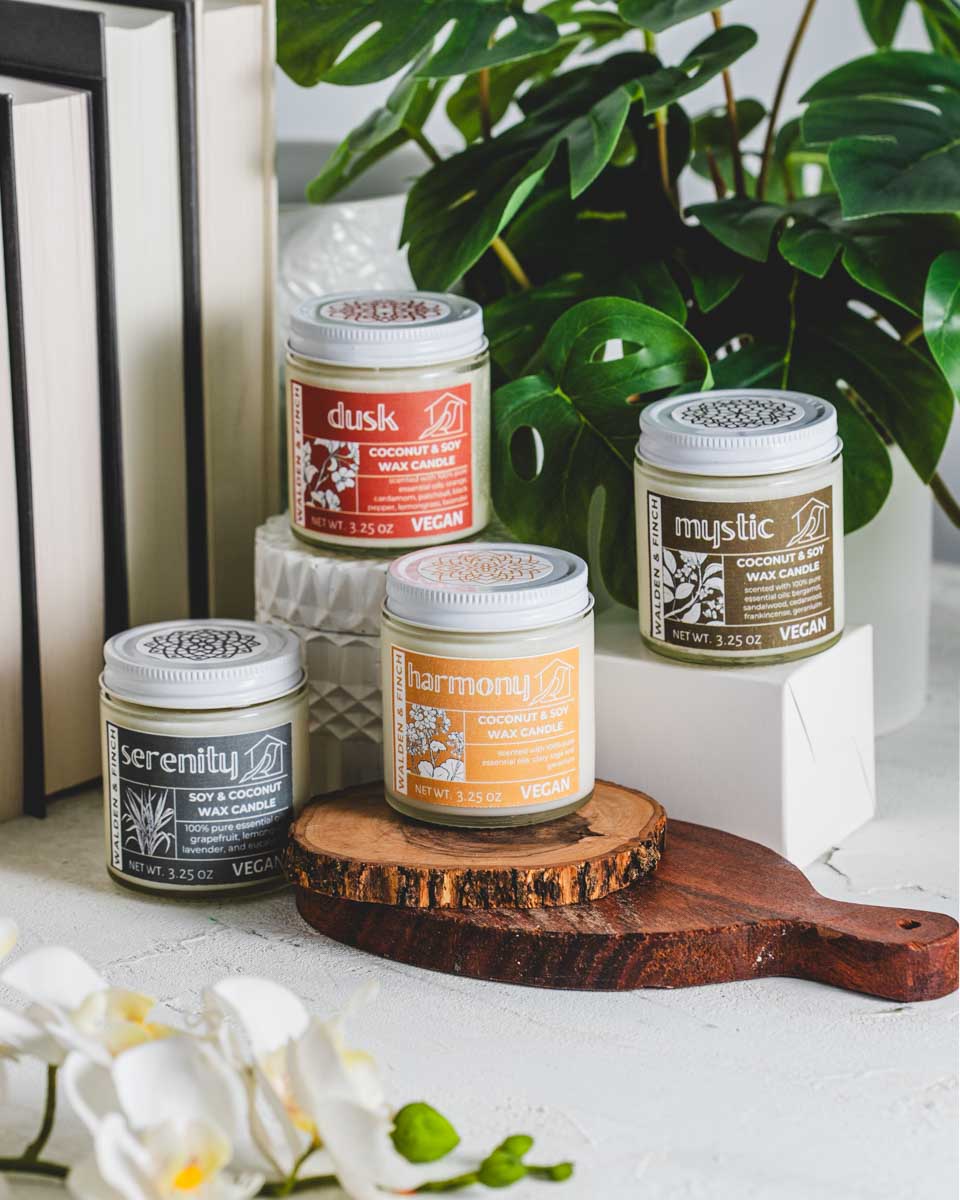 Four vegan candles labeled 'Serenity,' 'Dusk,' 'Harmony,' and 'Mystic' arranged on a wooden stand and table.
