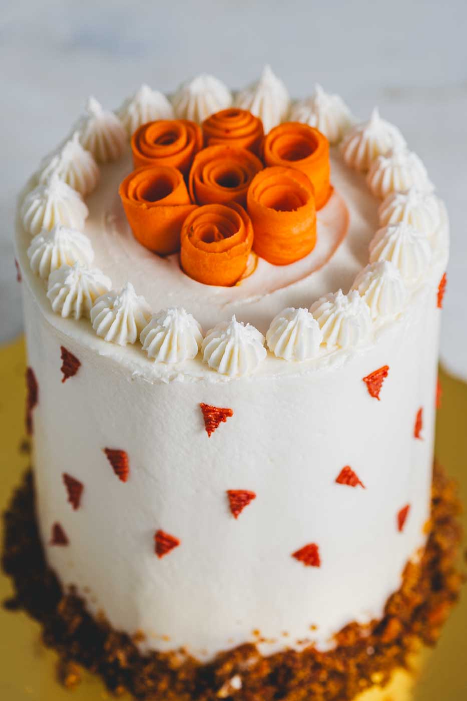 Carrot cake with decorative white frosting and a garnish of carrot "flowers". Around the sides there is decorative orange icing "carots" and the bottom is rolled in a crumb coating