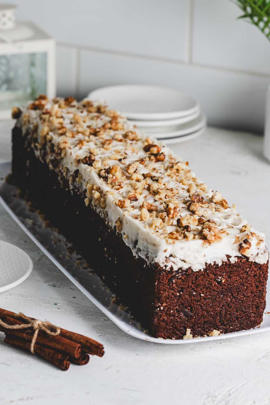 Carrot walnut loaf topped with creamy frosting and crushed walnuts, displayed on a white platter with cinnamon sticks.
