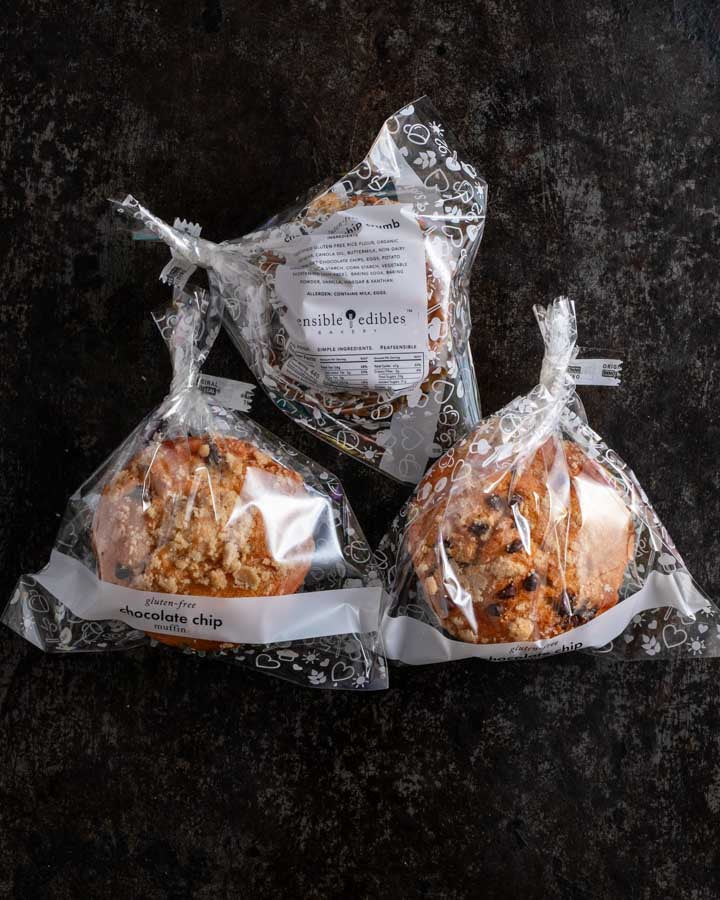 Three packaged chocolate chip muffins with crumb topping from Sensible Edibles, wrapped in clear plastic with white labels, on a dark rustic surface.