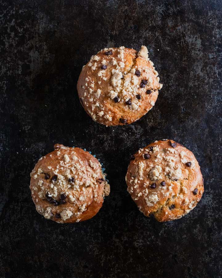 Three chocolate chip muffins with a crumb topping, arranged in a triangular pattern on a dark rustic surface.