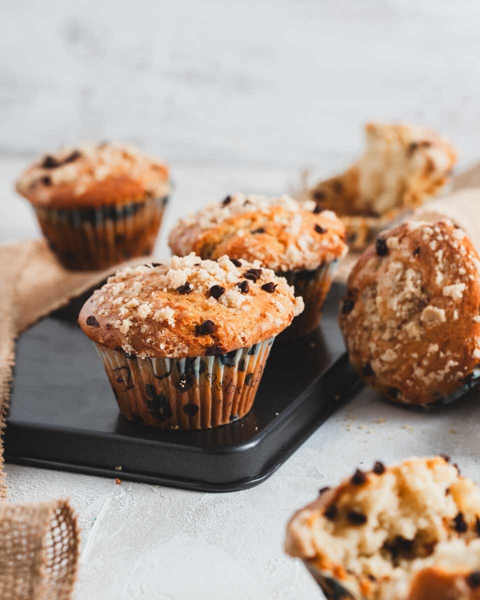 Chocolate chip muffins with a crumb topping, arranged on a black tray with additional muffins in the background, showcasing their golden-brown texture.