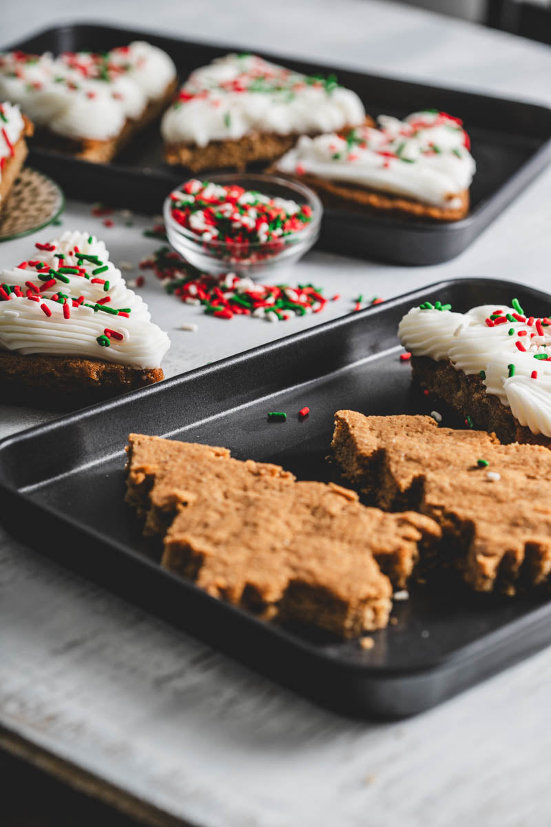 Christmas tree-shaped cookies with white frosting and red and green sprinkles on black trays, with a bowl of sprinkles in the background.