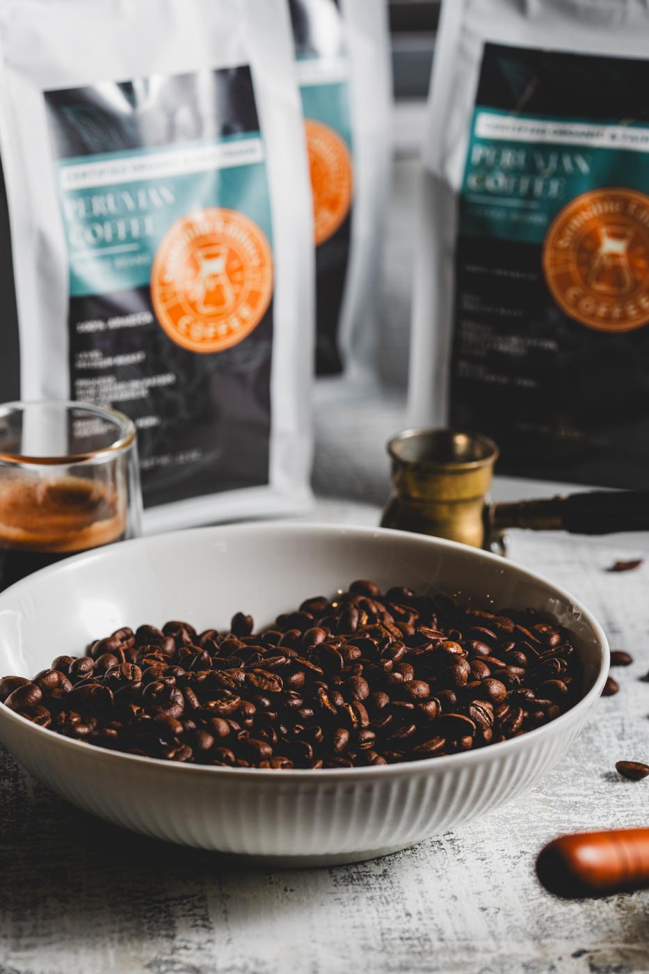A bowl of roasted coffee beans with bags of coffee behind it and a cup of coffee and coffee maker nearby