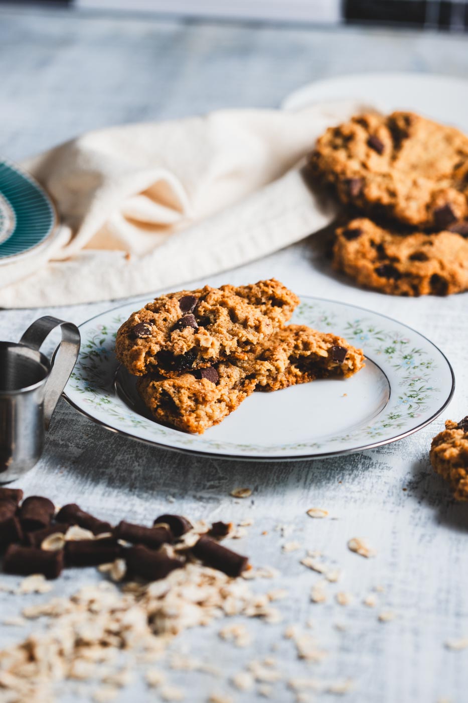 Oatmeal chocolate chip cookie broken open on a plate to show chocolate pieces and soft texture, with more cookies, oats, and chocolate pieces around