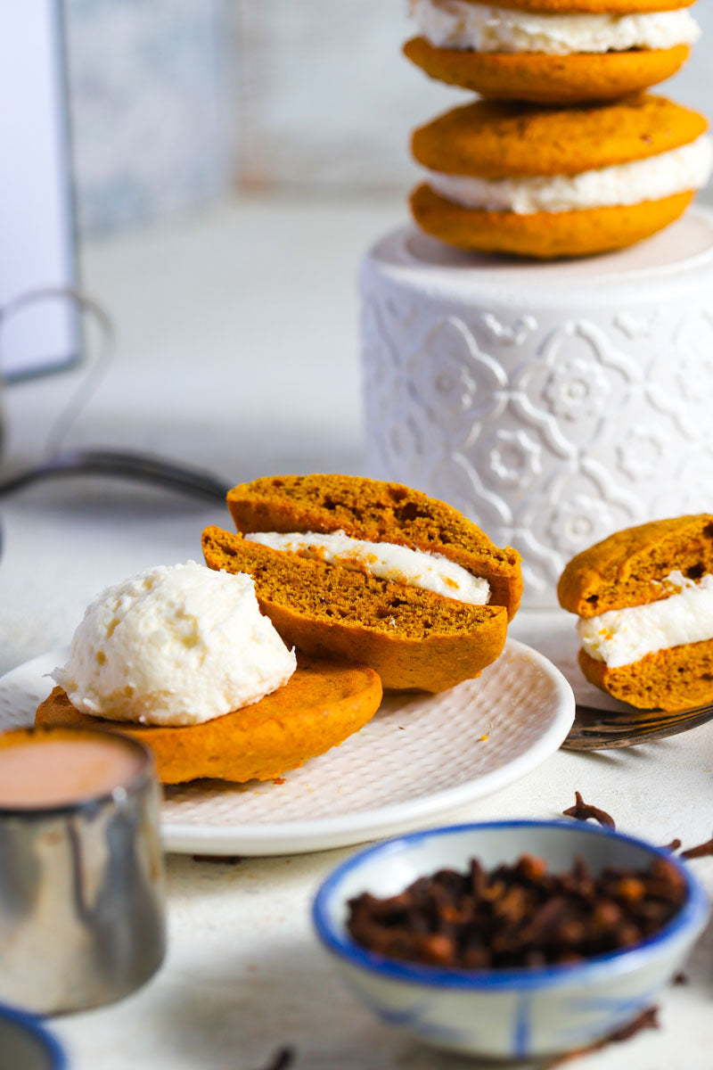 Pumpkin spice whoopies on and around  white plates with spices and coffee