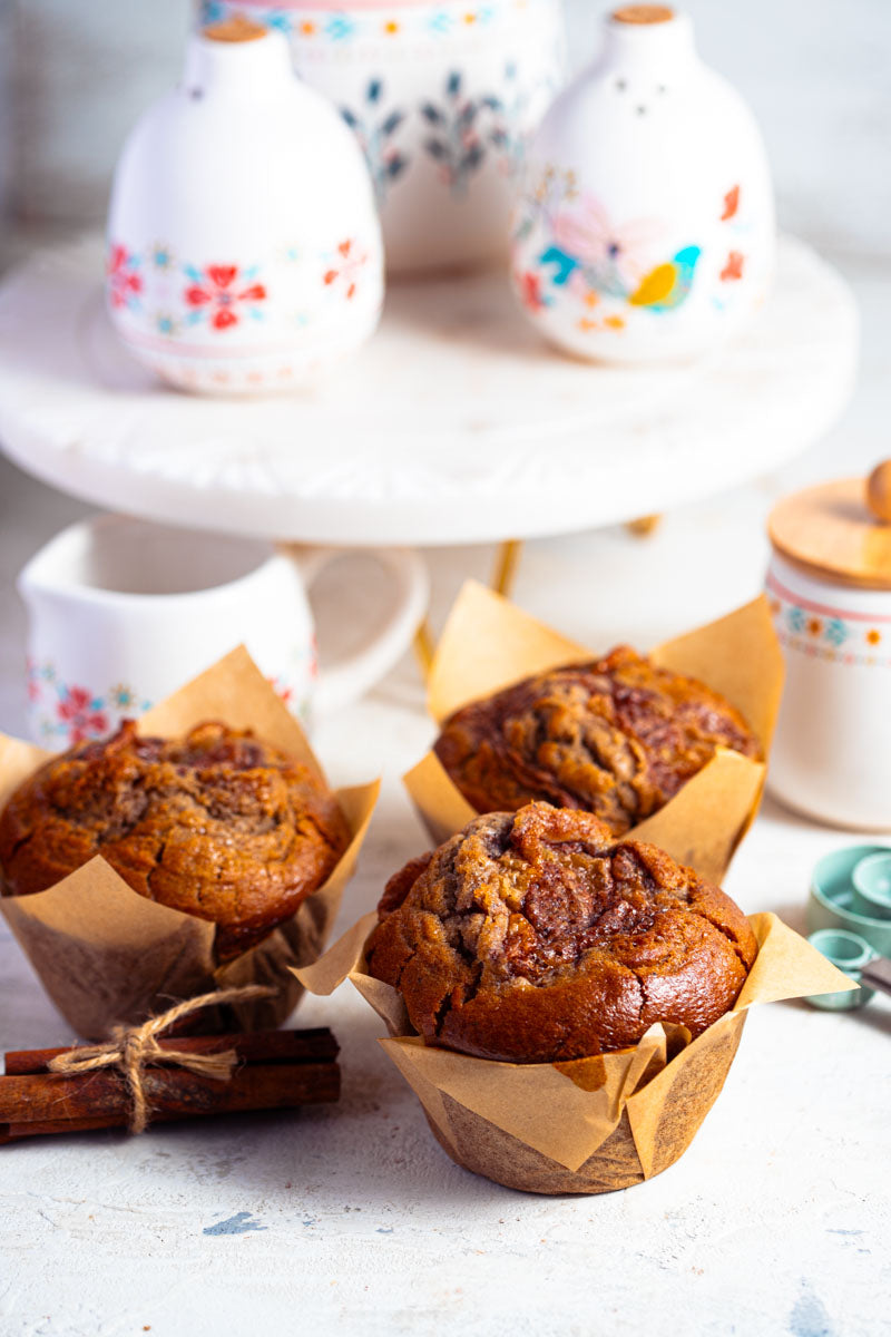 Three apple pie muffins in brown paper on a light surface with cinnamon sticks and ceramic kitchen items
