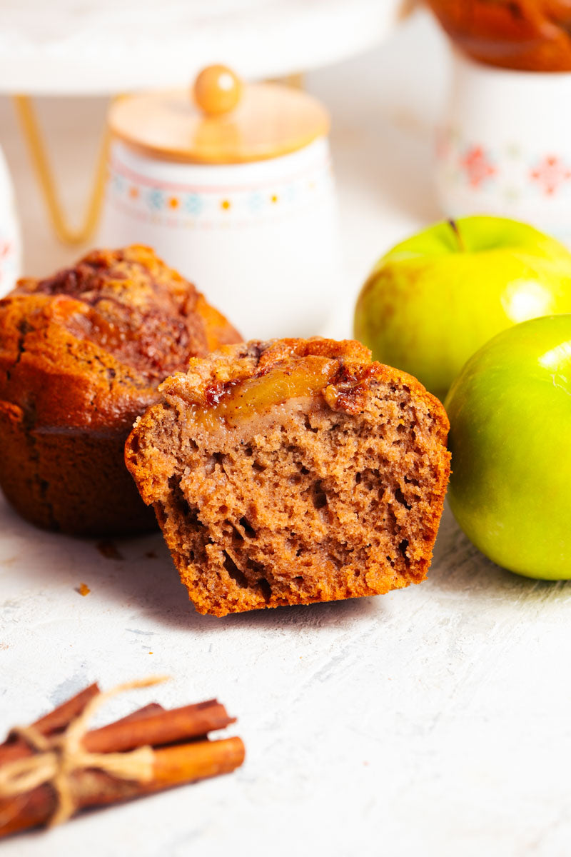 Apple pie muffin cut in half, surrounded by green apples and cinnamon sticks on a white surface.