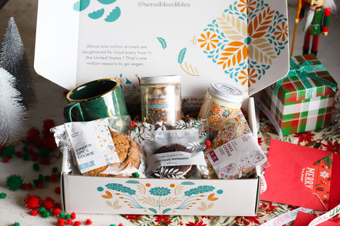 Holiday gift box with a mug, hot chocolate jar, candle, ginger spice cookie, peppermint whoopie, and christmas cookie surrounded by christmas decorations.
