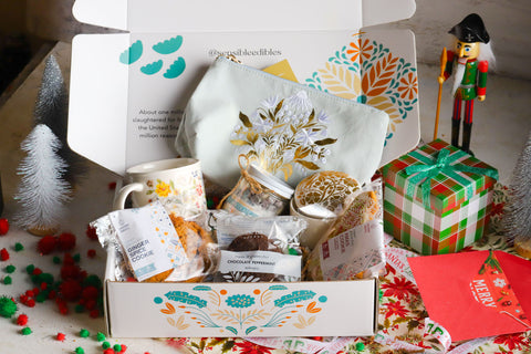 Holiday gift box with mug, hot chocolate jar, embroidered bag, ceramic trinket box, ginger spice cookie, peppermint whoopie, and frosted christmas cookie surrounded by christmas decorations.