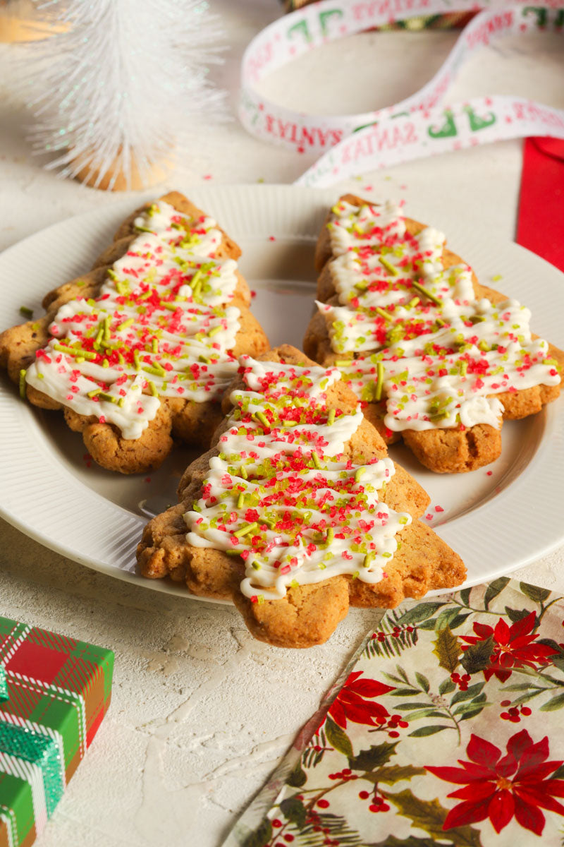 Three frosted christmas cookies shaped like trees with green and red sprinkles on a white plate surrounded by christmas decorations