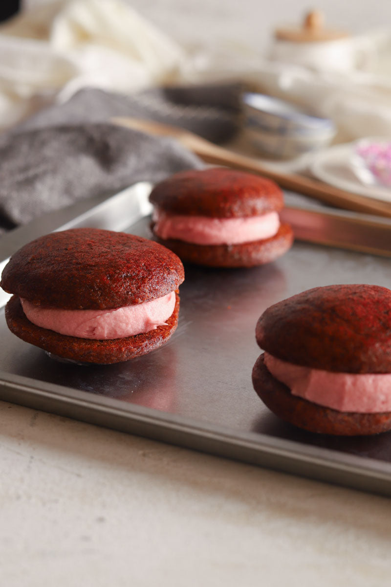 Red velvet vegan gluten free whoopie pies with pink filling on a baking tray.
