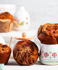 Apple pie muffins on a white surface with a floral mug and apples and cinnamon sticks in the background