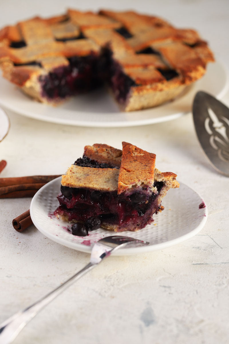 Slice of blueberry pie on a plate with a whole pie in the background