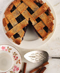 A blueberry lattice pie seen from above with a slice taken out. Around it is a cake server, cinnamon sticks, and a plate.