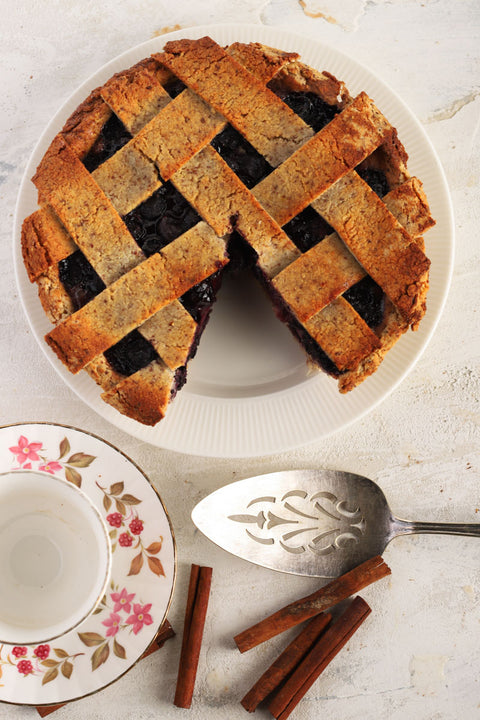 A blueberry lattice pie seen from above with a slice taken out. Around it is a cake server, cinnamon sticks, and a plate.