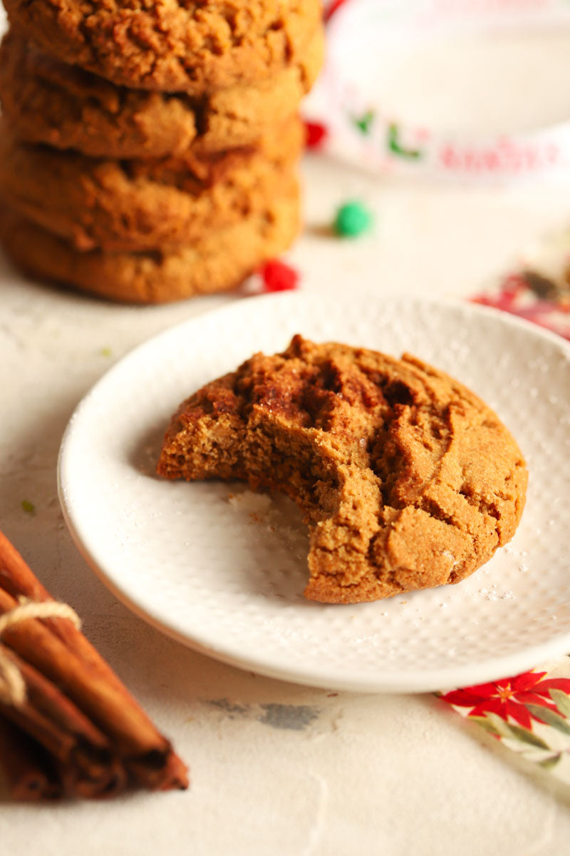 Gingerbread spice cookie with a bite taken out on a white plate with a cinnamon stick and Christmas decorations in the background.