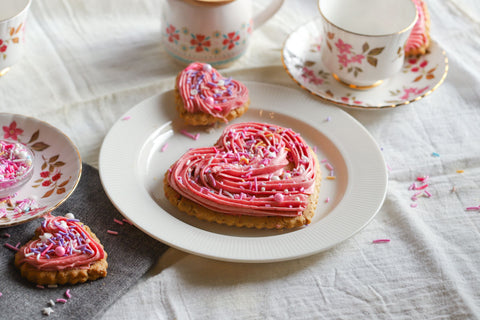 Heart-shaped cookies with pink frosting and sprinkles on a white plate, surrounded by floral teacups and saucers.