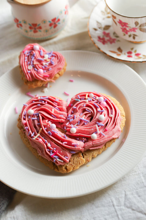 Heart-shaped cookies with pink frosting and sprinkles on a white plate, with floral teacups in the background.