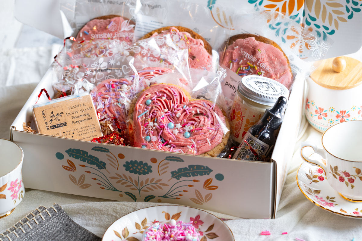 Valentine's gift box of sweetheart cookies,pretty in pink cookies, a candle, soap, and body oil with tea cupson a table.