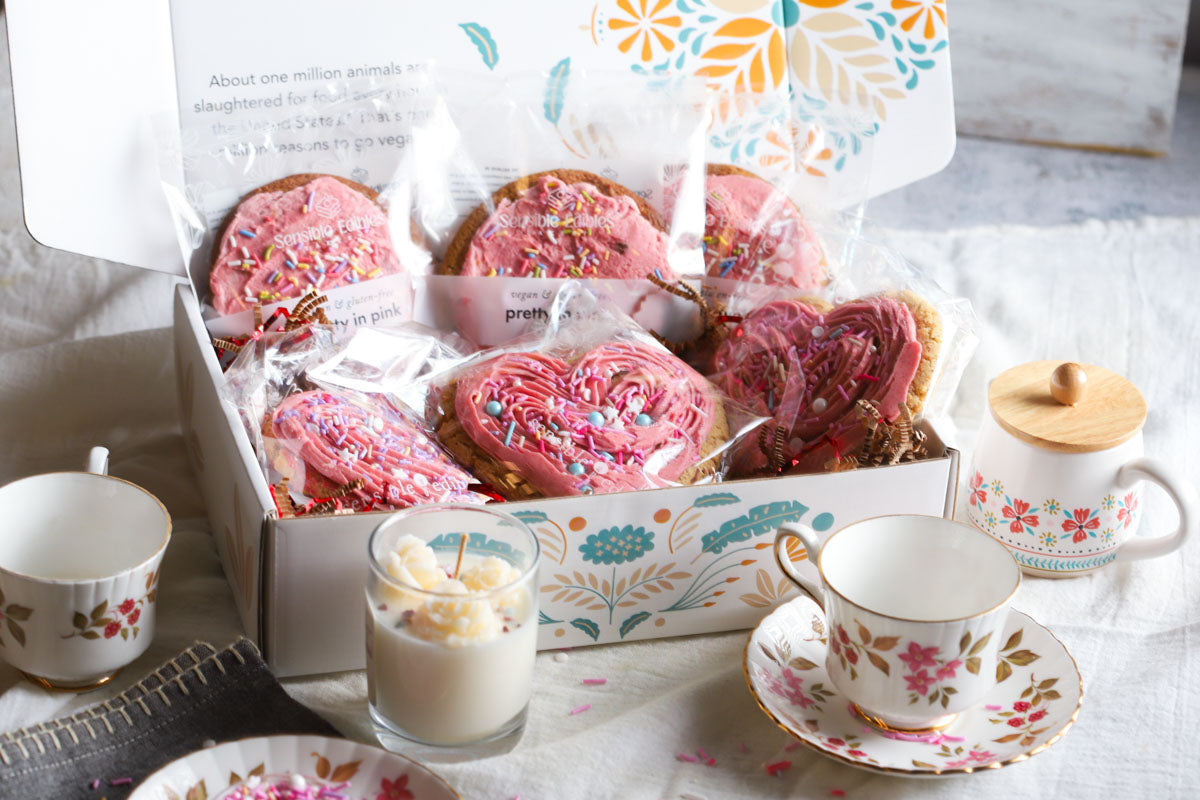 Valentine's gift box of sweetheart cookies and pretty in pink cookies with tea cups and a candle on a table.