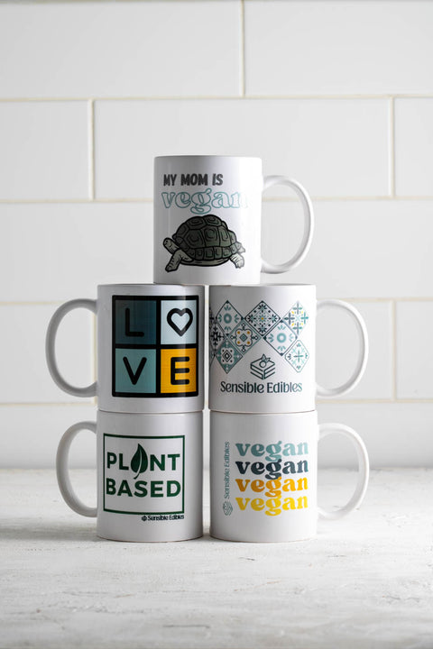 Stack of six Sensible Edibles mugs with various designs, including: 'My Mom is Vegan' with a turtle, 'LOVE', 'Plant Based', 'Sensible Edibles' with a geometric pattern, and 'Vegan Vegan Vegan'.