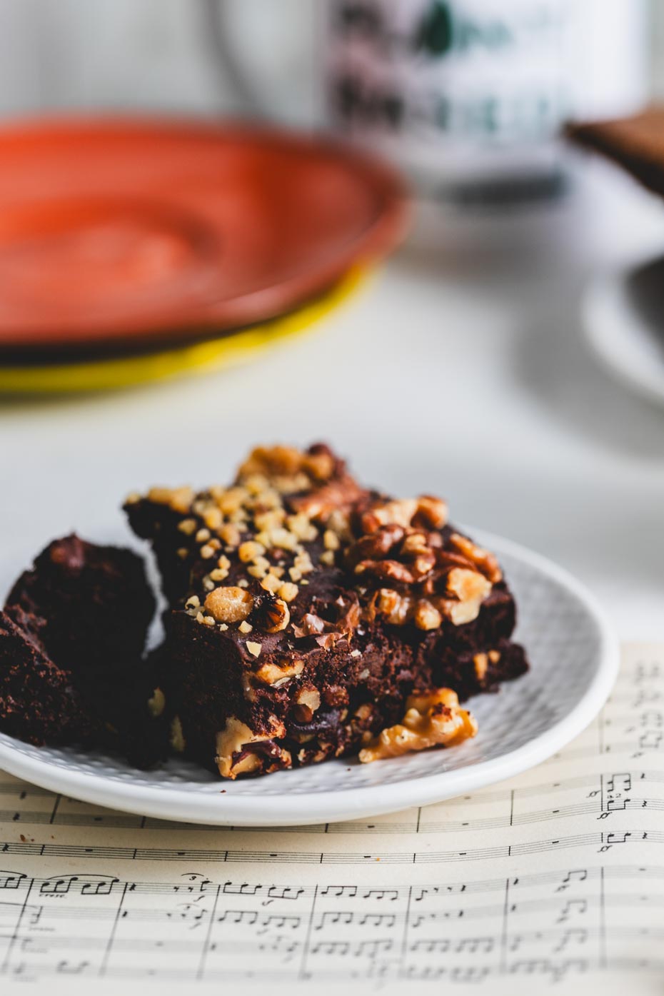Close-up of a walnut brownie topped with chopped nuts on a white plate, placed on top of sheet music.