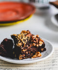 Close-up of a walnut brownie topped with chopped nuts on a white plate, placed on top of sheet music.