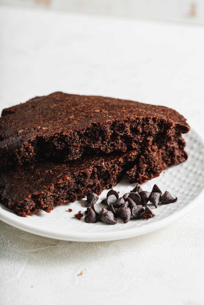 A broken milk chocolate cookie on a white plate, with a few chocolate chips scattered beside it, showing the soft and chewy interior.