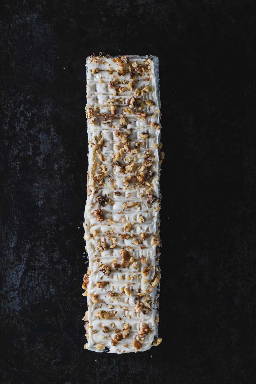 Top view of a carrot walnut loaf topped with creamy frosting and crushed walnuts, showcasing intricate frosting details on a dark surface.