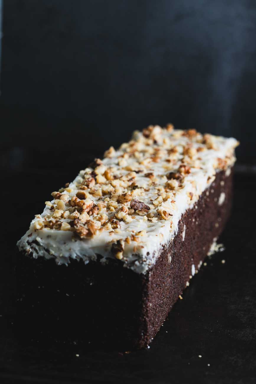 Carrot walnut loaf topped with creamy frosting and crushed walnuts, displayed on a dark surface.