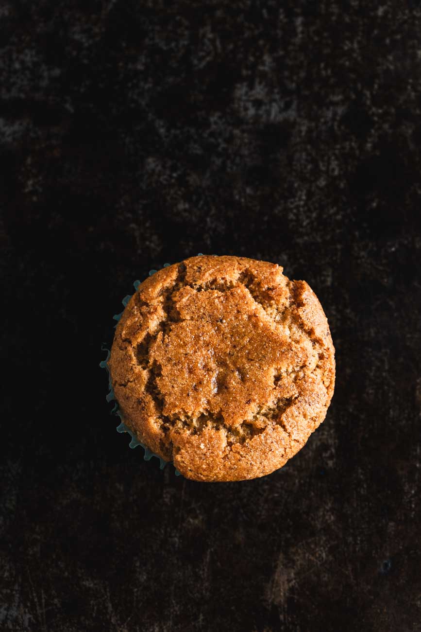 A close-up of a single golden-brown muffin on a dark background, showcasing its cracked top and textured surface.