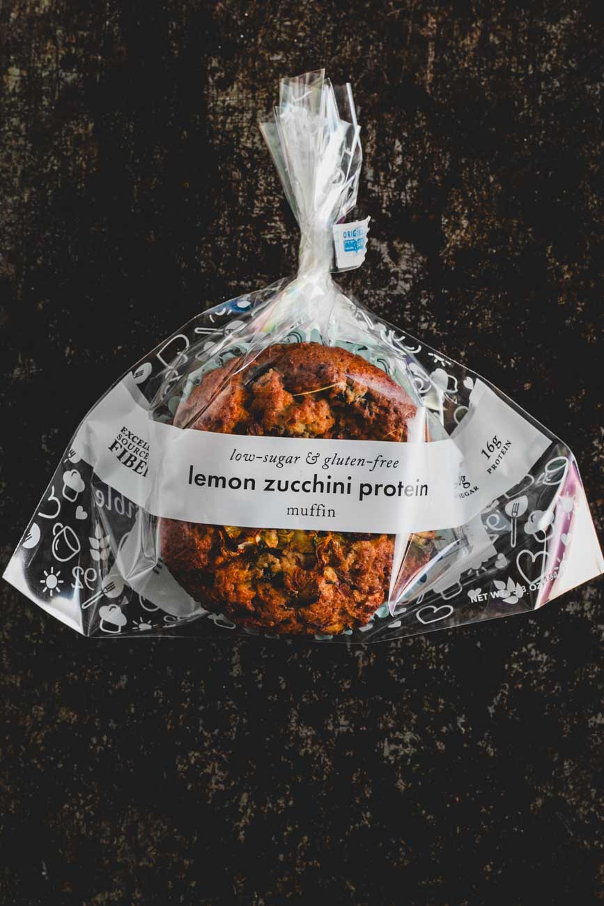 A packaged lemon zucchini protein muffin in a clear plastic bag with a white label on a dark background. The label reads 'low-sugar & gluten-free lemon zucchini protein muffin.