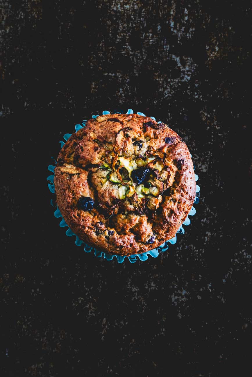 Top view of a single lemon zucchini protein muffin in a blue paper liner on a dark background. The muffin is golden brown with visible zucchini shreds and blueberries.