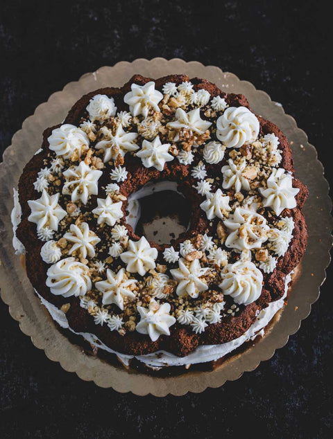 Large carrot walnut bundt cake with creamy frosting and crushed walnuts on a scalloped-edge cake board, featuring intricate frosting designs